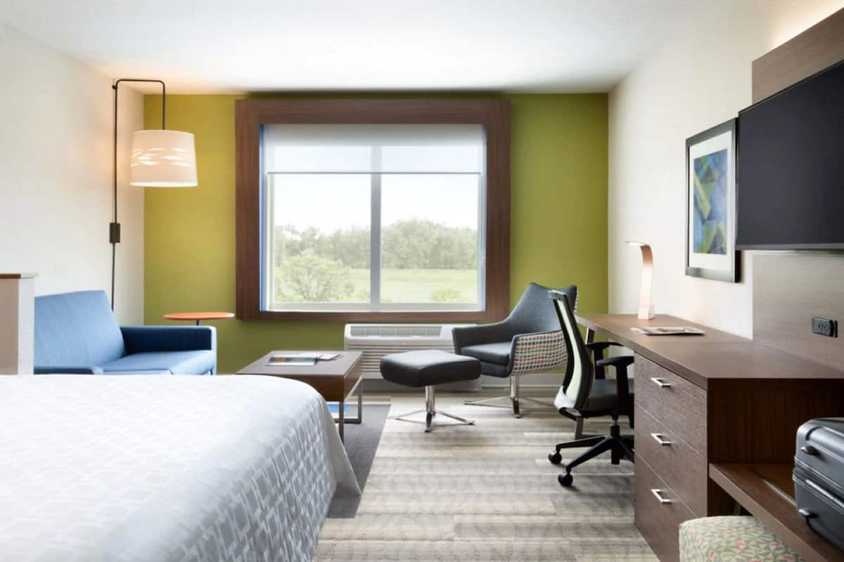 A hotel room with a bed, a desk, a chair, a lamp, and a window with a view of greenery.