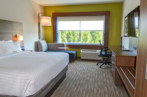 A hotel room with a large bed, a desk, a chair, a sofa, a window with a view of trees, and a flat-screen TV. - Holiday Inn Express & Suites Raleigh Airport - Brier Creek by IHG (Raleigh)