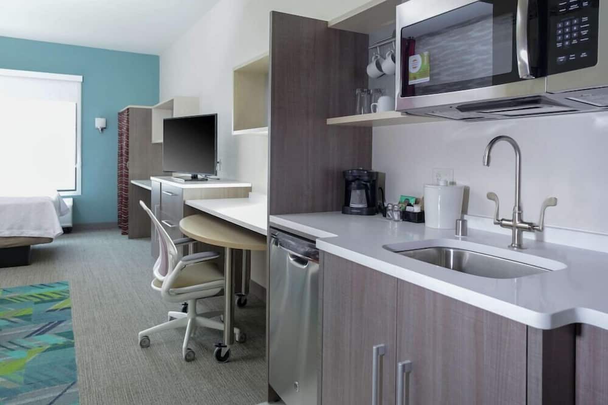 A modern hotel room with a kitchenette, a desk, a chair, a bed, and a bathroom.