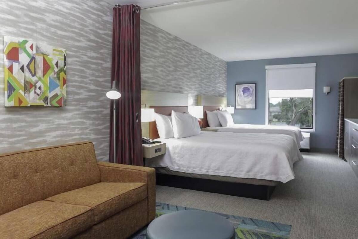 A hotel room with a bed, a sofa, a nightstand, a lamp, and a window.