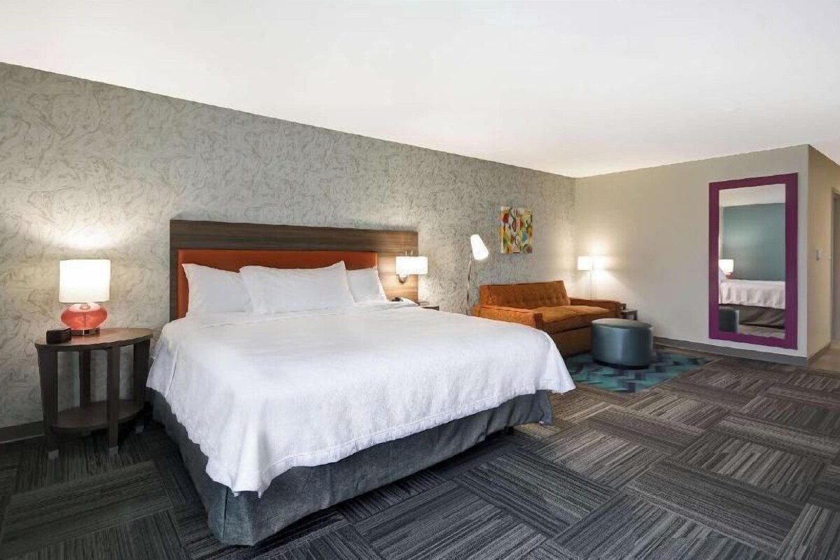 Une chambre d’hôtel avec un grand lit, deux lampes de chevet, une chaise, une petite table ronde et un miroir.