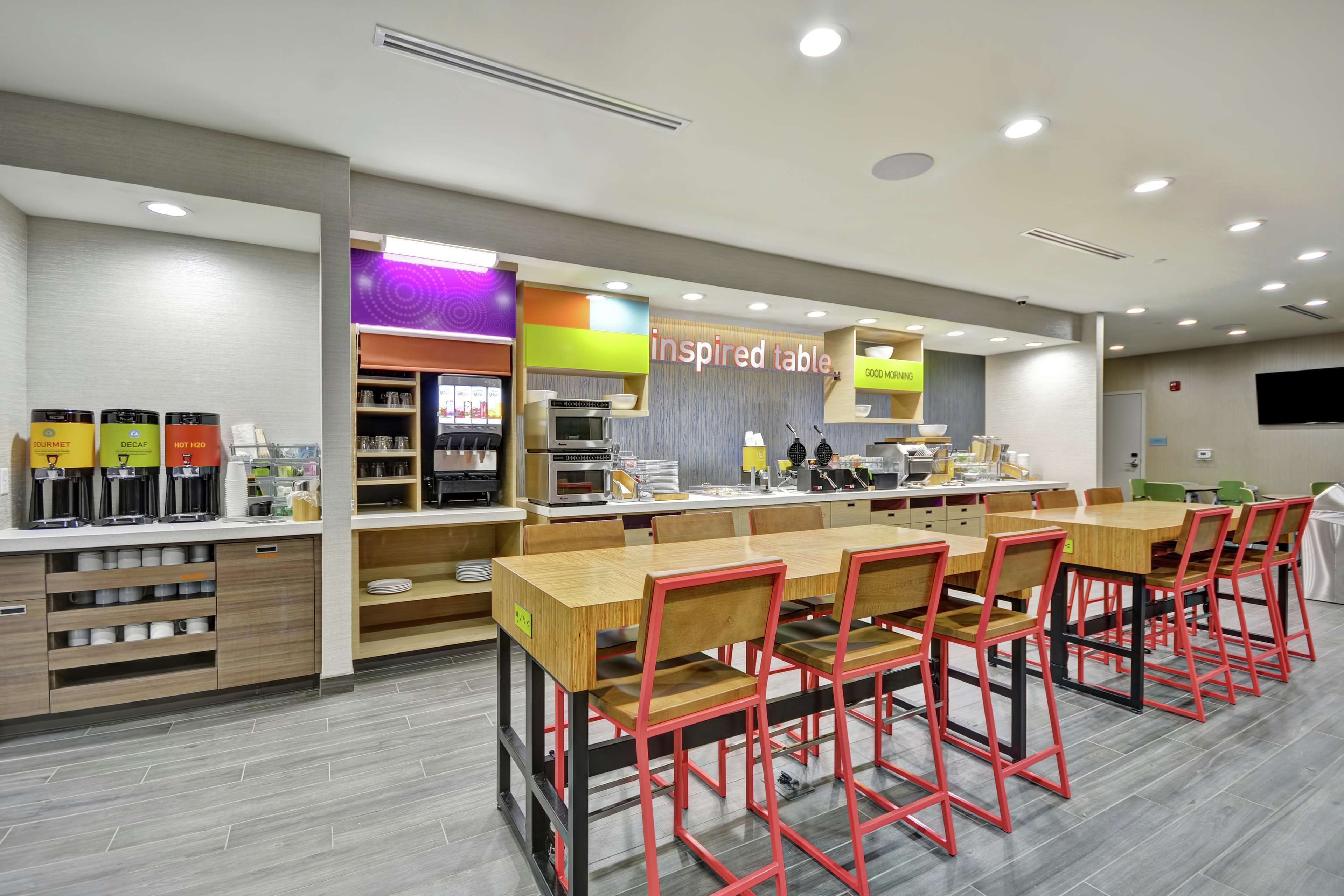 A modern cafe with a long wooden table and red chairs, a coffee machine, and a sign that reads "inspired table."