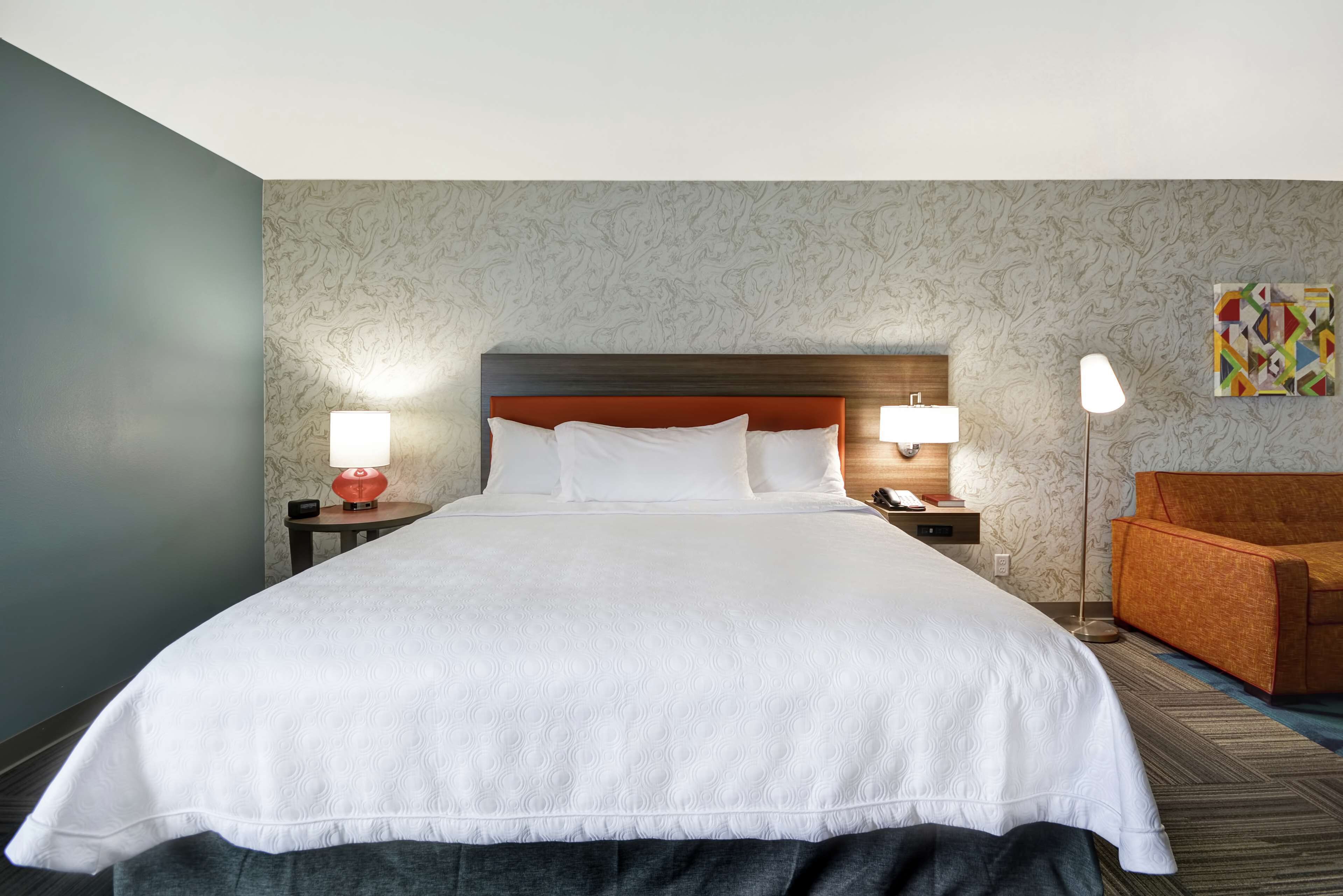 Un grand lit avec une tête de lit en bois, deux tables de chevet avec des lampes, une chaise et un mur décoré d’un motif.