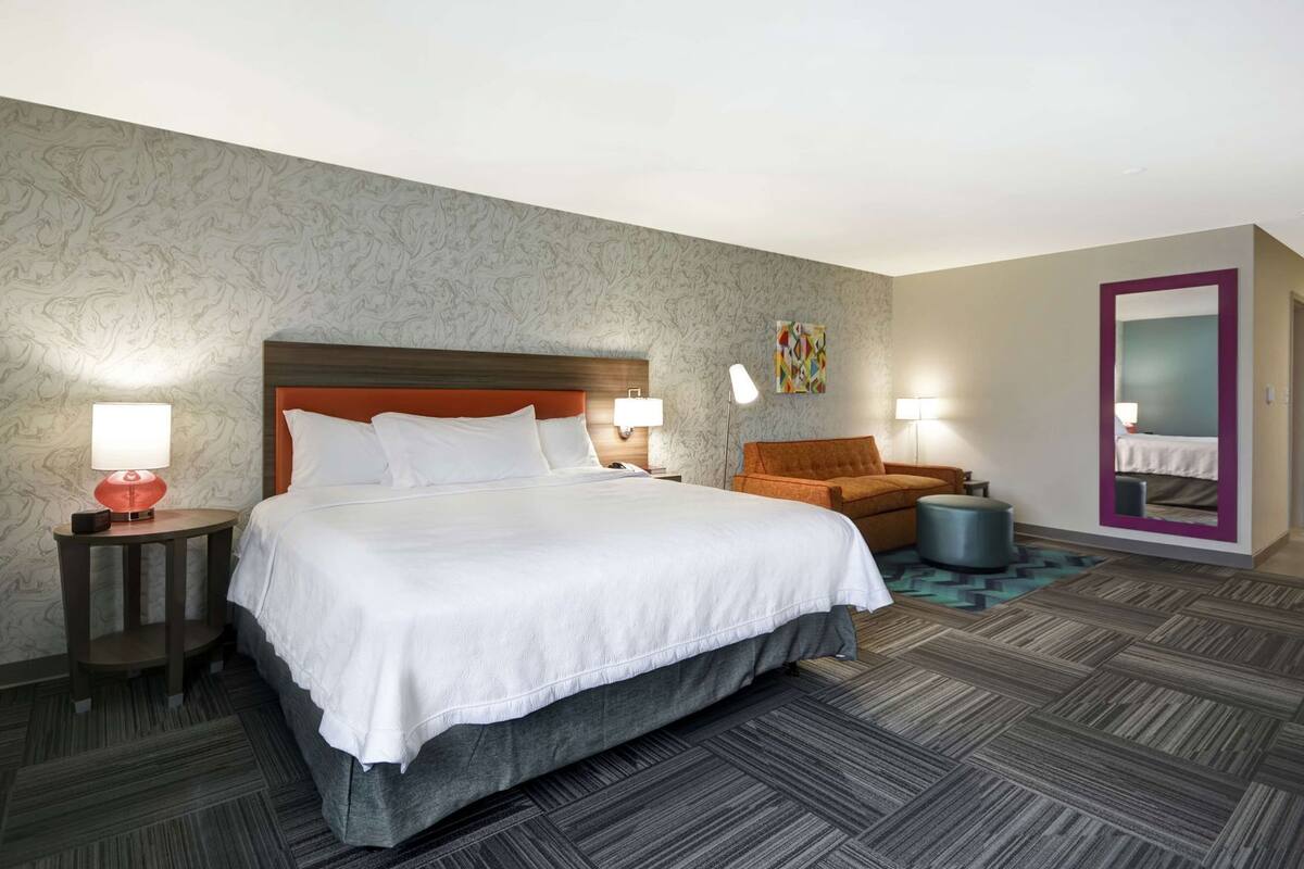 Una habitación de hotel con una cama grande, dos mesitas de noche, un sofá, un otoman redondo, un espejo y una alfombra estampada.