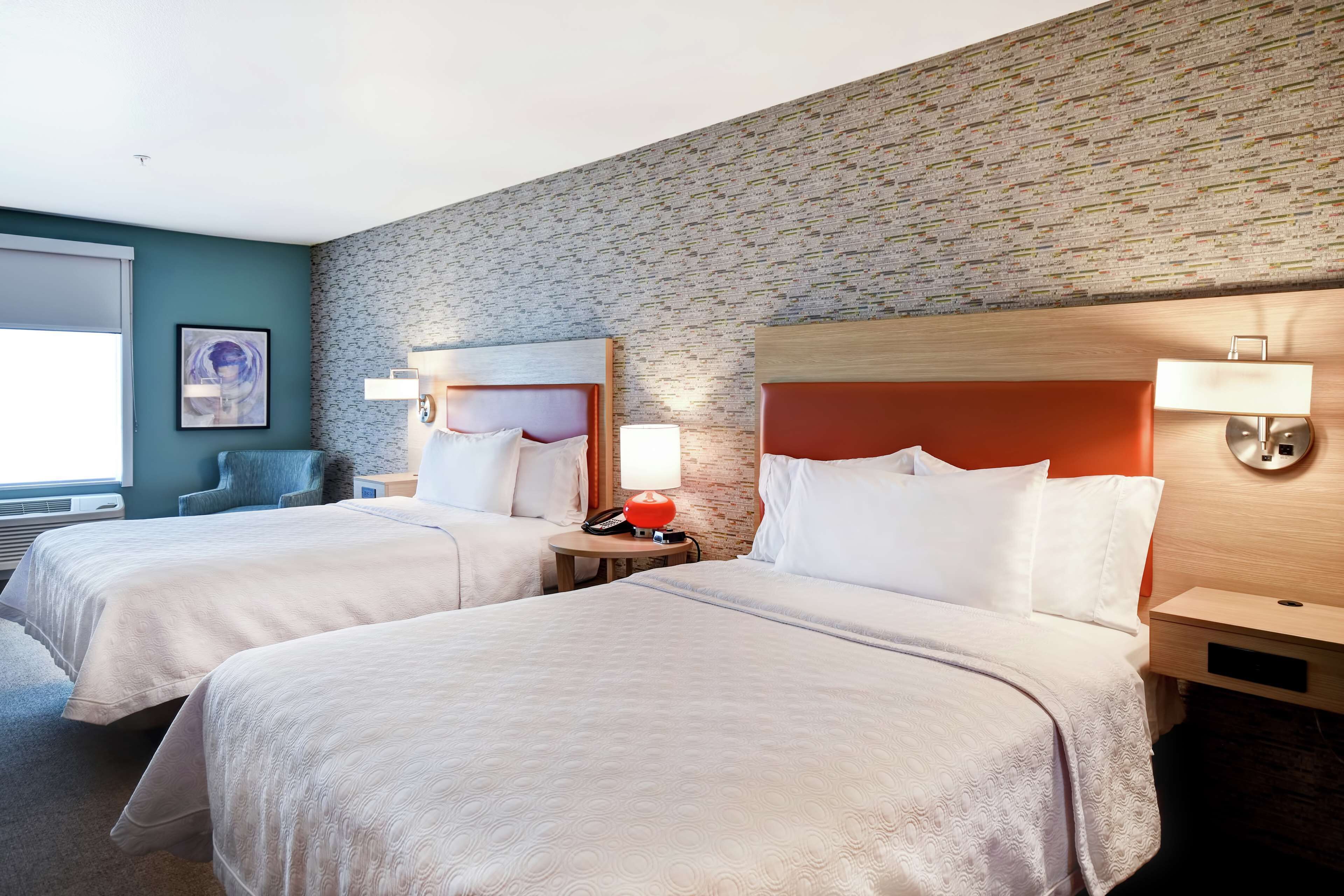 A hotel room with two beds, a brick-patterned wall, a framed artwork, and a window with blinds.