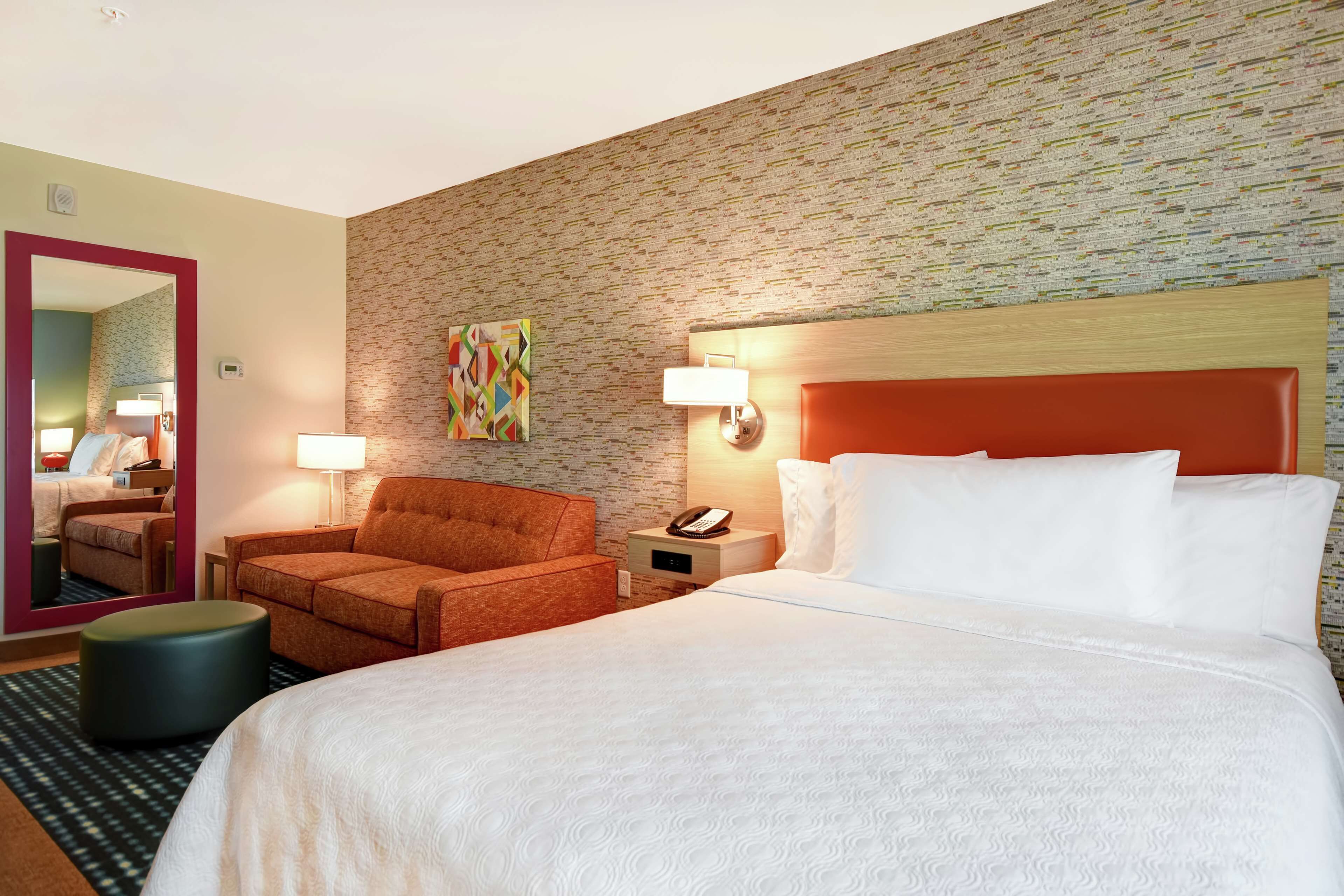 Quarto de hotel com uma cama grande, um sofá, uma mesinha, uma mesa de cabeceira com abajur, um telefone e um espelho.