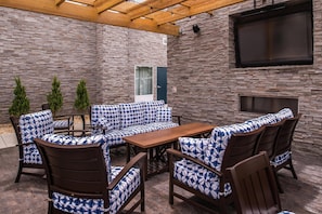 Un patio al aire libre con muro de piedra, techo de madera, televisor montado en la pared y zona de estar con cojines estampados y sillas de madera.