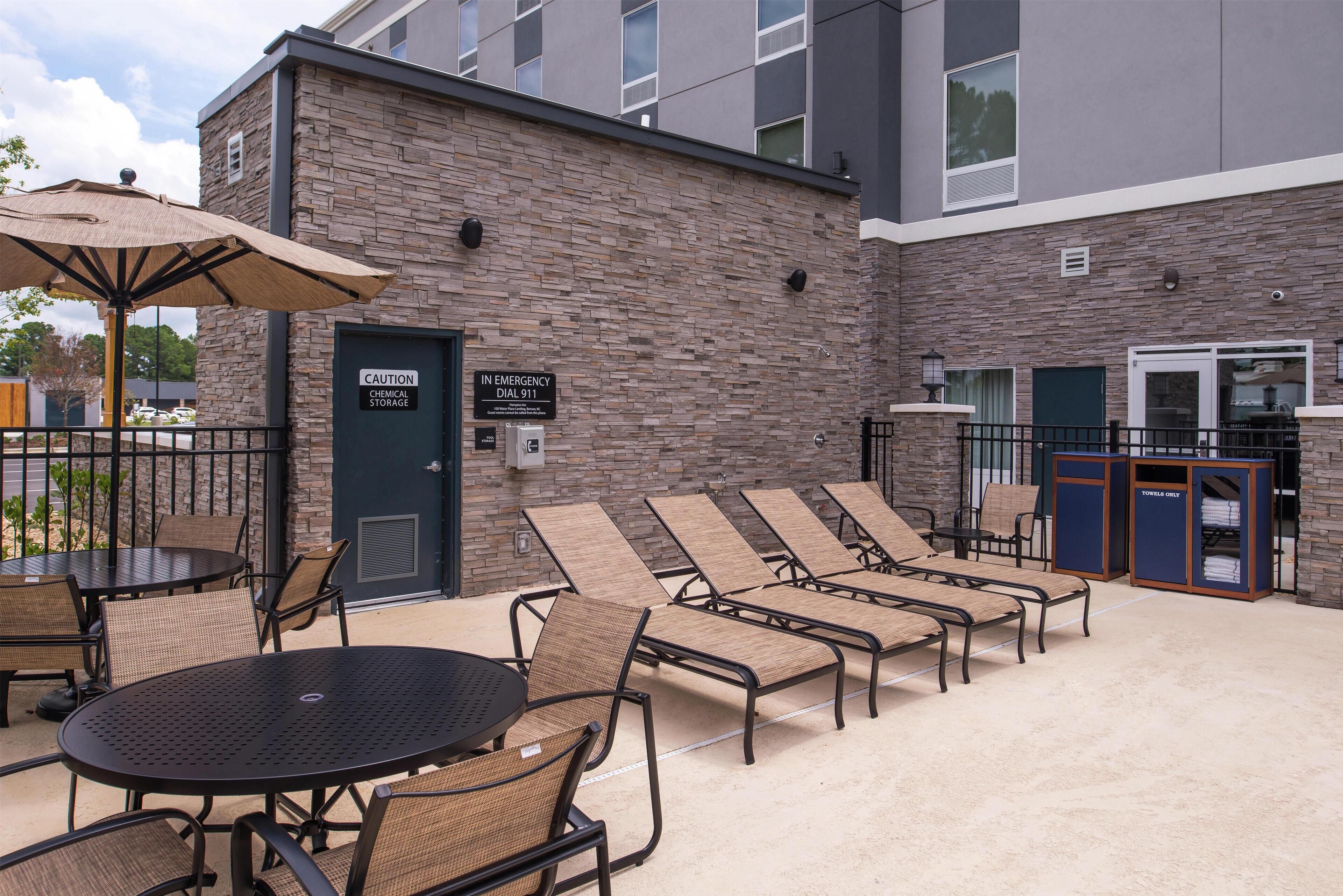 A patio area with lounge chairs, a round table, and a covered seating area. There is a sign indicating an emergency dial 911.