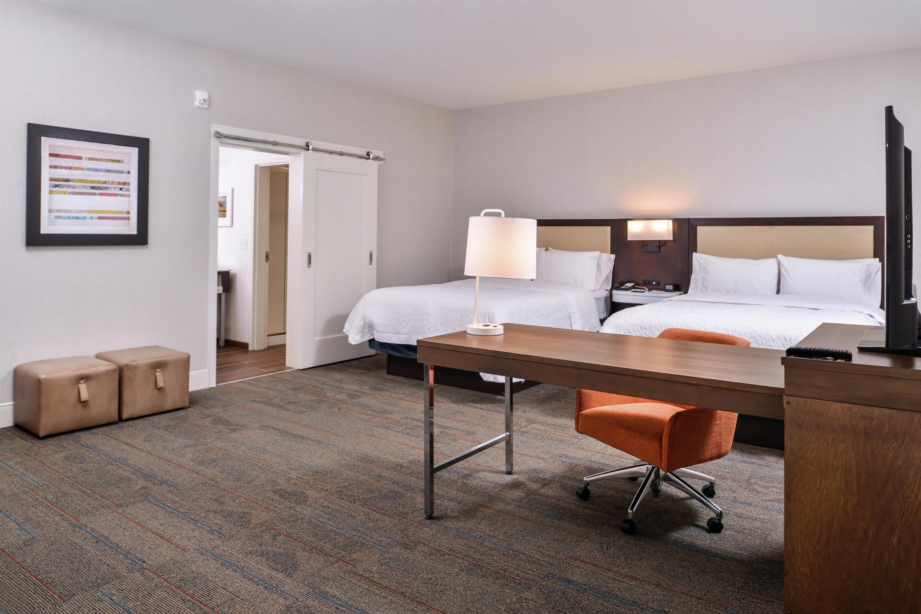 A hotel room with two beds, a desk, an orange chair, and a TV.