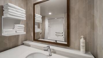 A bathroom with a large mirror, a sink, and a shower. There are white towels stacked on a shelf.