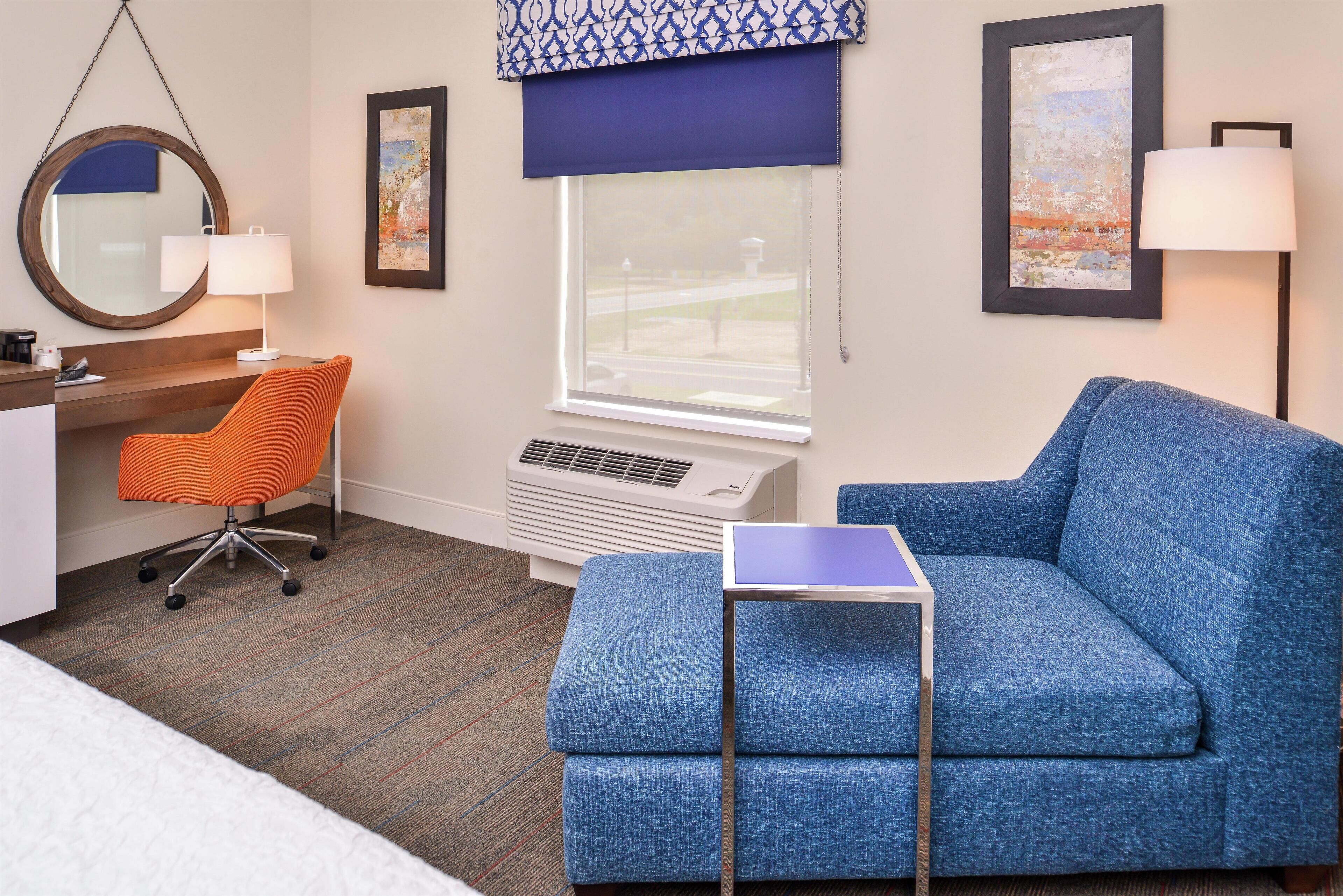 A hotel room with a blue sofa, an orange office chair, a small table, a desk, a large window with a view, and a wall-mounted air conditioner.