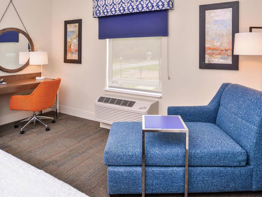 A hotel room with a blue sofa, an orange office chair, a small table, a desk, a large window with a view, and a wall-mounted air conditioner.
