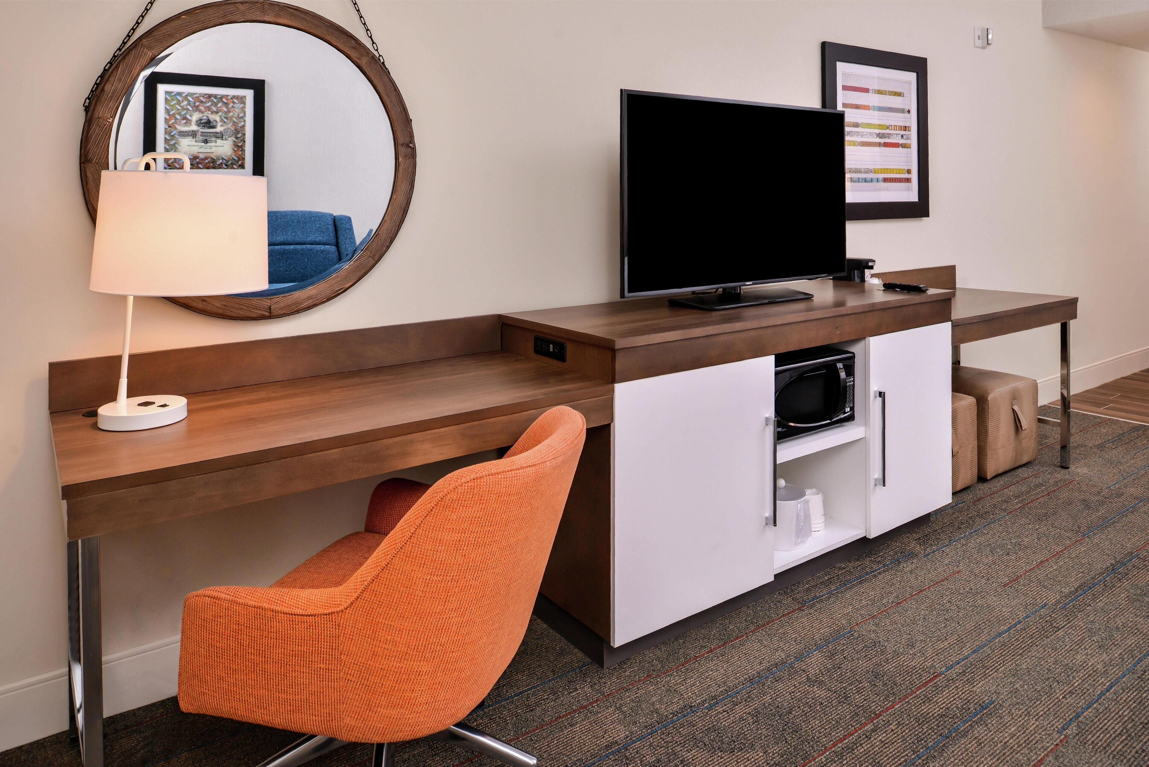 A hotel room with a wooden desk, a television, a mirror, and a single orange chair.