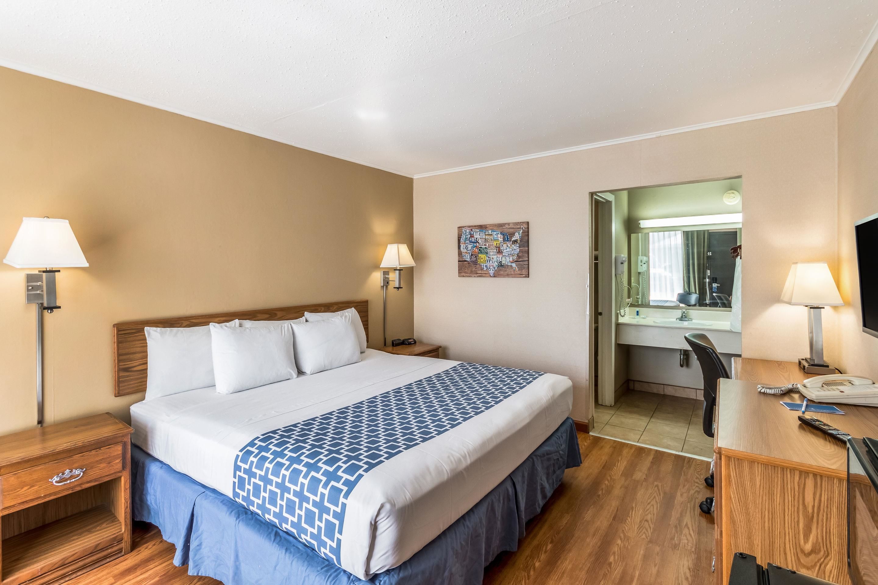 Une chambre d’hôtel avec un grand lit, des tables de chevet avec des lampes, un bureau avec une chaise et une salle de bain visible à travers une porte ouverte.