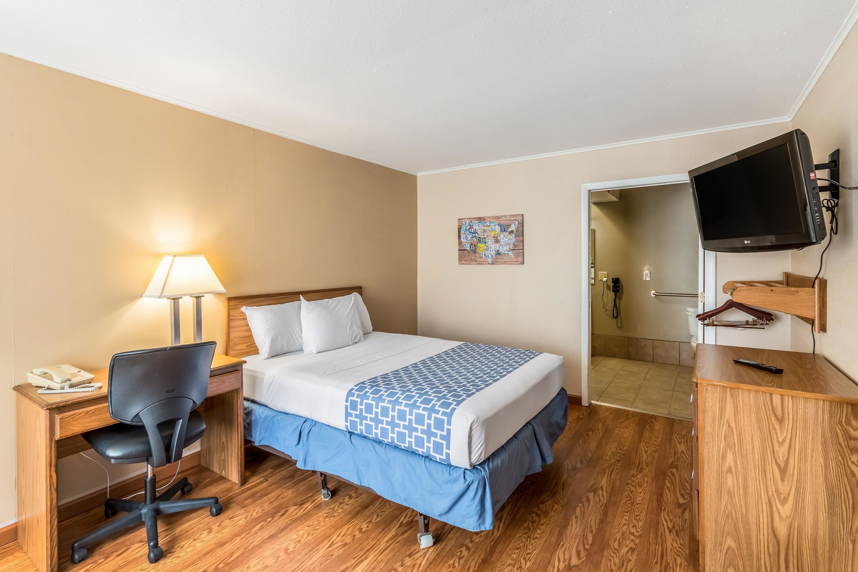 A hotel room with a bed, desk, chair, lamp, TV, and a wooden dresser.