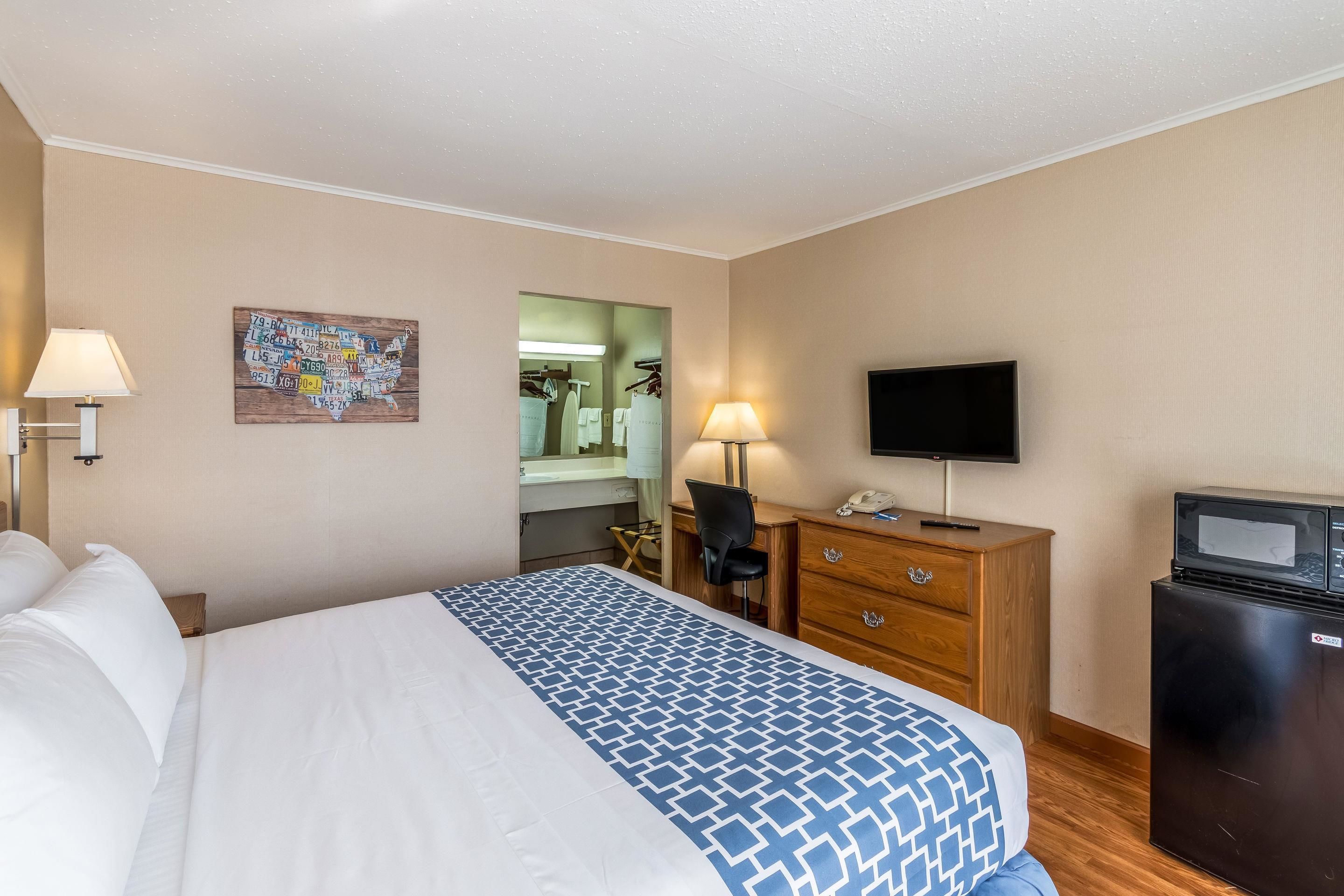 Una habitación de hotel con cama, escritorio con silla, televisor, refrigerador y espejo.