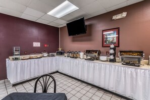 Montaje de bufé con una larga mesa cubierta con tela blanca, diferentes estaciones de comida, un microondas y un televisor de pantalla plana montado en la pared.