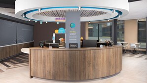 A modern hotel reception area with a wooden reception desk, a digital screen, and a sign indicating a 24/7 market.