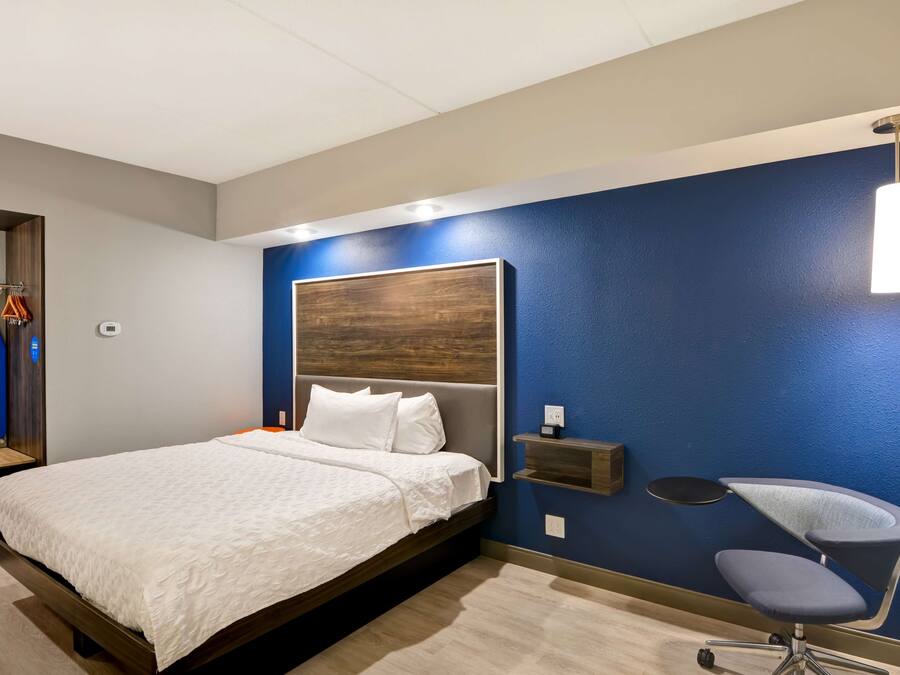 Quarto de hotel moderno com uma cama grande, uma escrivaninha com cadeira, uma prateleira na parede e uma luminária pendente.