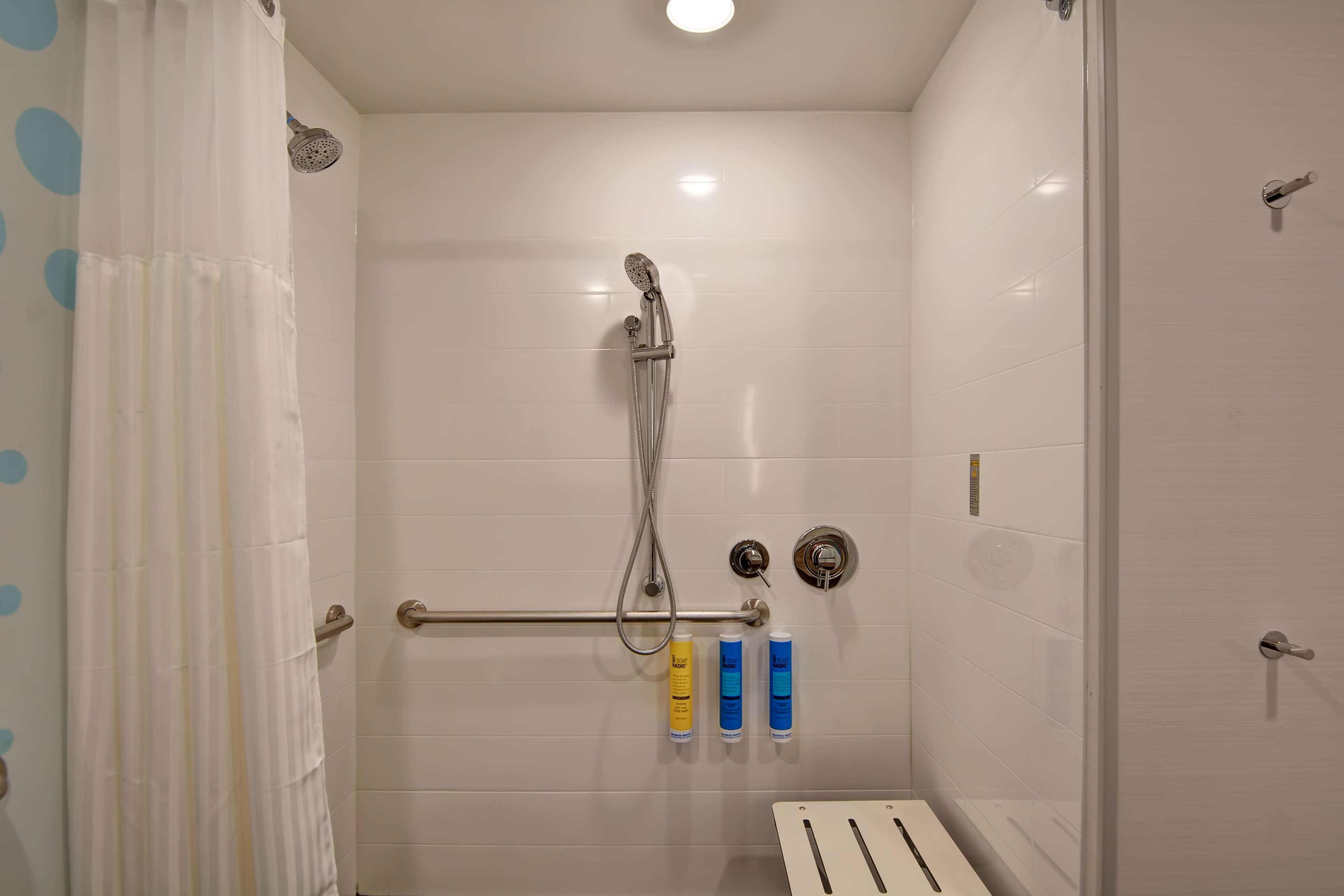 A walk-in shower with a handheld showerhead, a handheld showerhead mounted on the wall, and a handrail. There are three bottles of shampoo and conditioner on the wall.