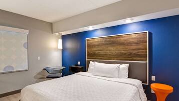 A modern hotel room with a large bed, a wooden headboard, a small desk, and a bright orange accent chair.