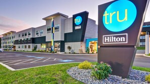 Un moderno edificio de hotel con un llamativo letrero 'tru Hilton', varios espacios de estacionamiento y una zona ajardinada bien cuidada.