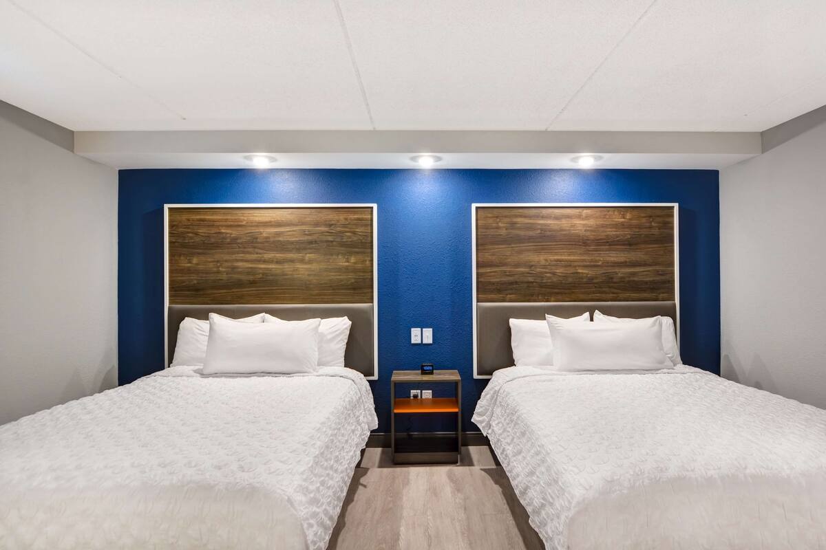 Habitación de hotel con dos camas, una pared decorada en azul, cabeceros de madera, ropa de cama blanca y una mesita de noche con un reloj.