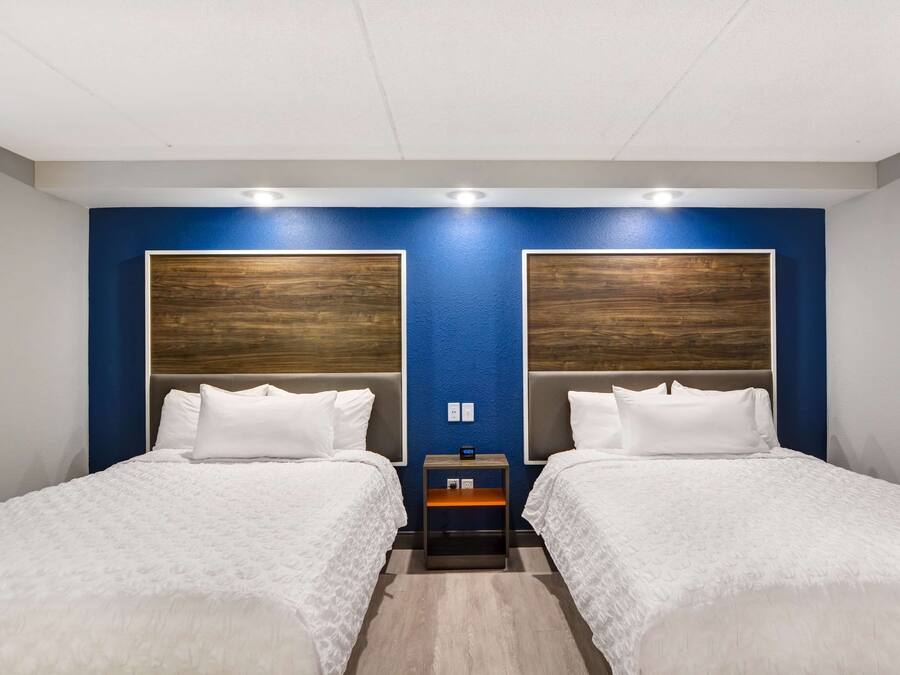 Camera d'albergo con due letti, una parete azzurra, testiere in legno, biancheria da letto bianca e un comodino con una sveglia.