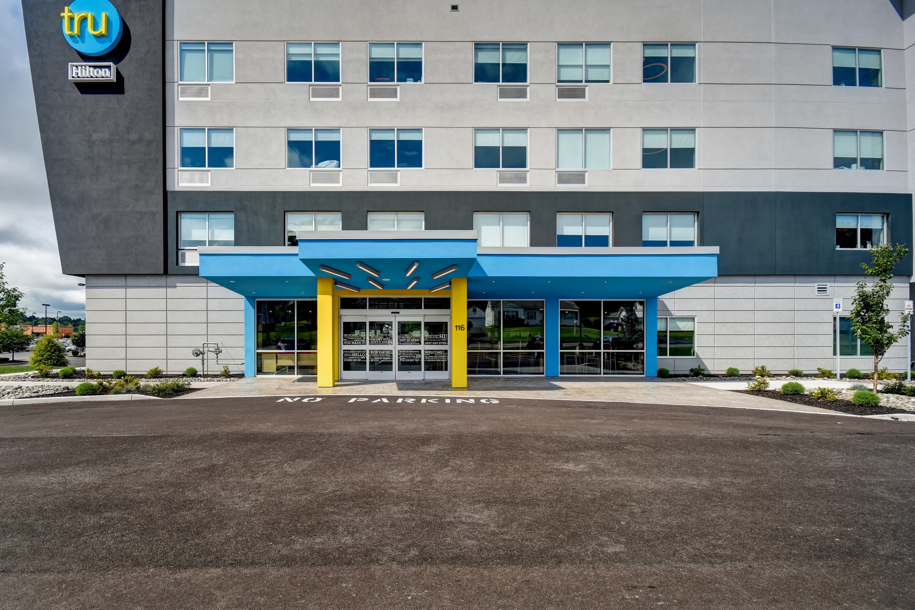Una entrada de hotel moderna con un toldo azul y amarillo, un área de estacionamiento señalizada como "PARKING", y el logotipo de Hilton.