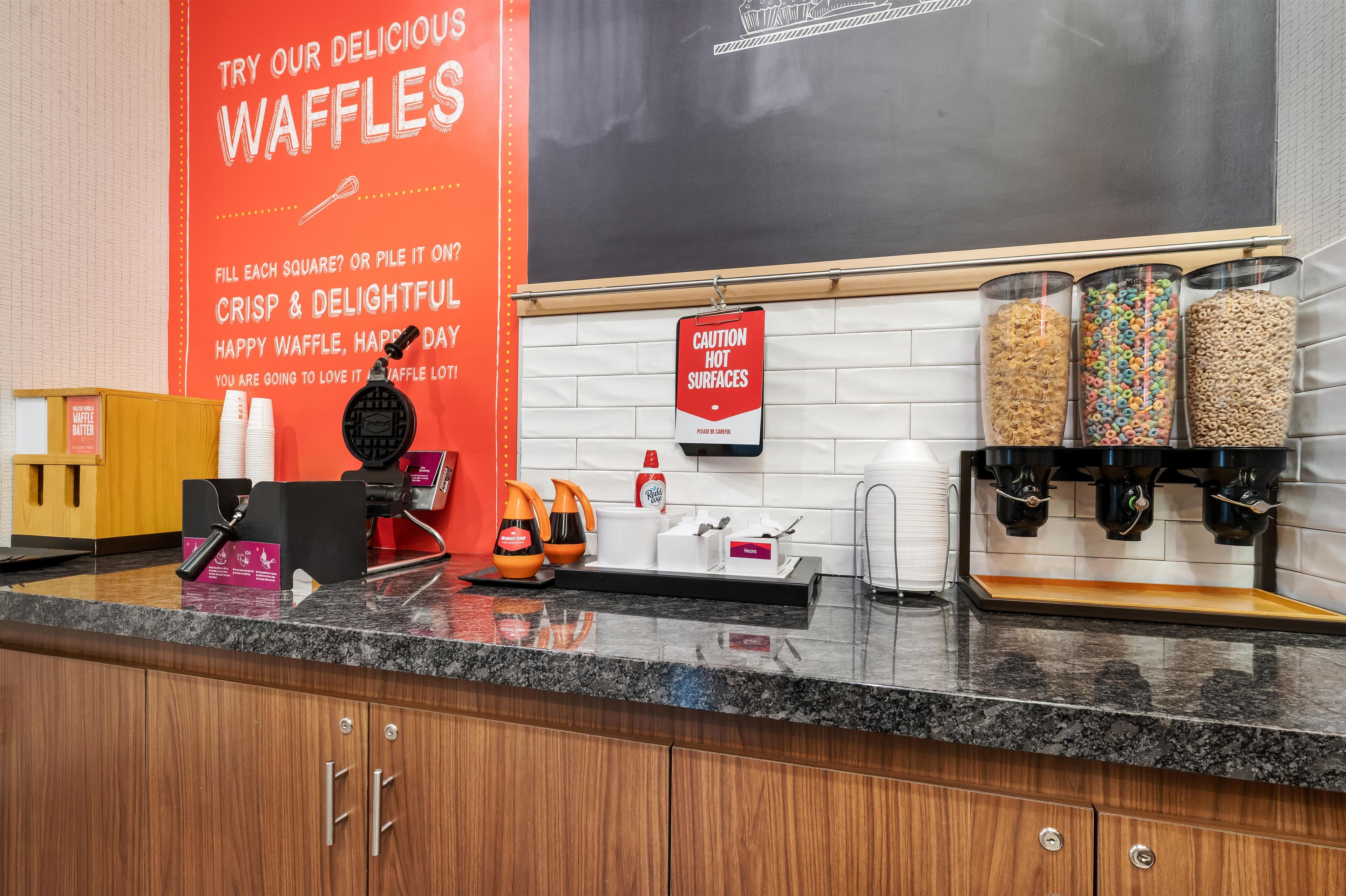 A kitchen counter with waffle-making equipment, a sign for waffles, and a caution sign for hot surfaces.