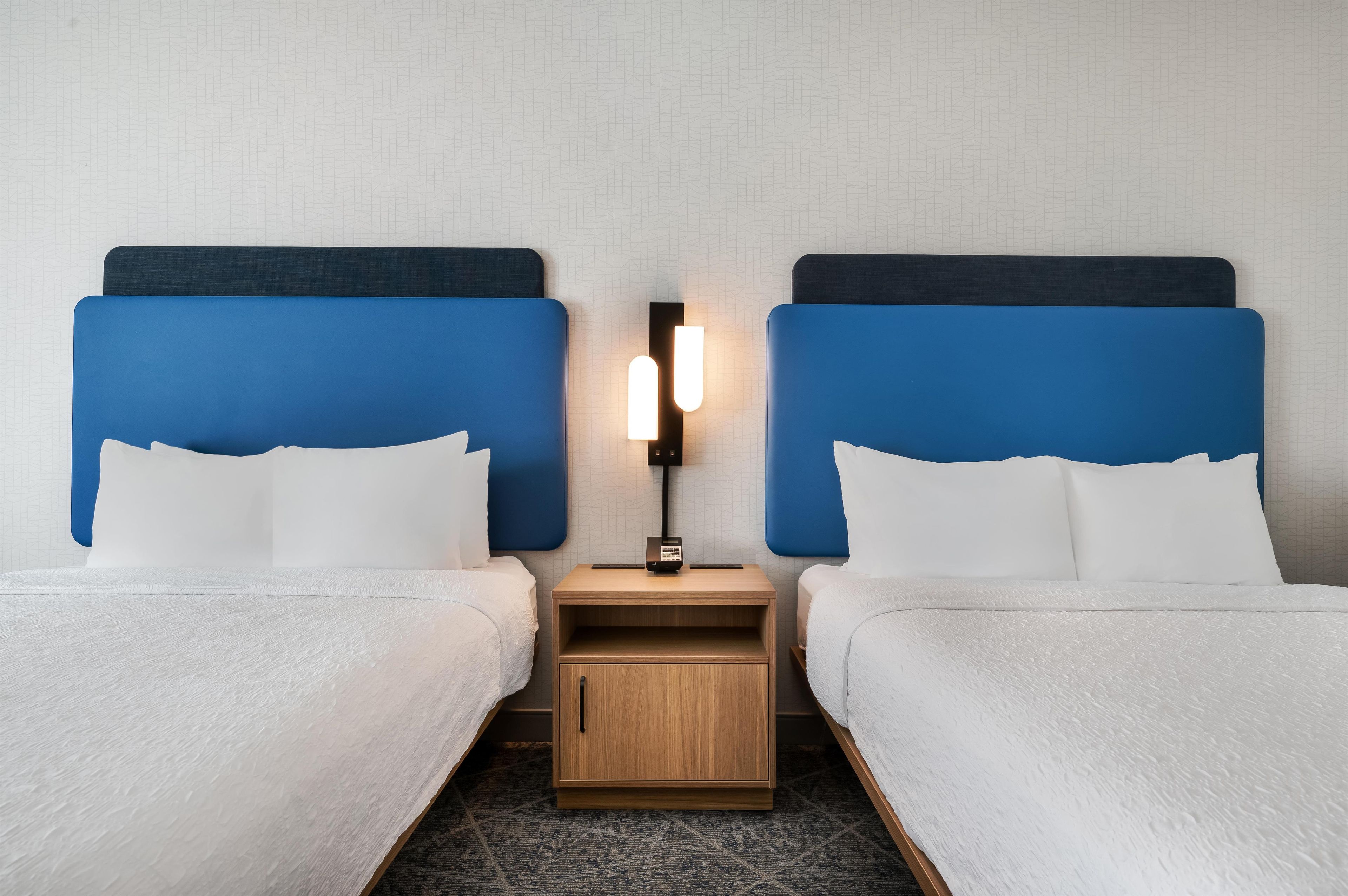 A hotel room with two beds, a nightstand, and a wall-mounted lamp.