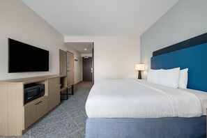 Habitación de hotel con cama, televisor, escritorio con microondas y puerta que da a un pasillo.
