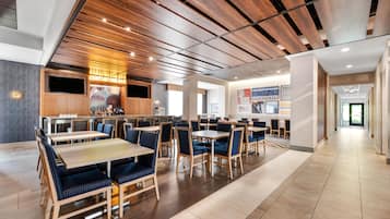 Un restaurante moderno con paneles de techo de madera, varias mesas de comedor con asientos de cubo acolchados en azul, y una zona de bar con una pantalla grande.