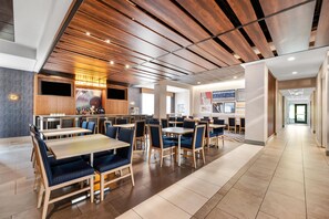 Ein modernes Restaurant mit Holzbodenpaneelen, mehreren Esstischen und blauen gepolsterten Sitzecken sowie einer Bar mit einem großen Bildschirm.