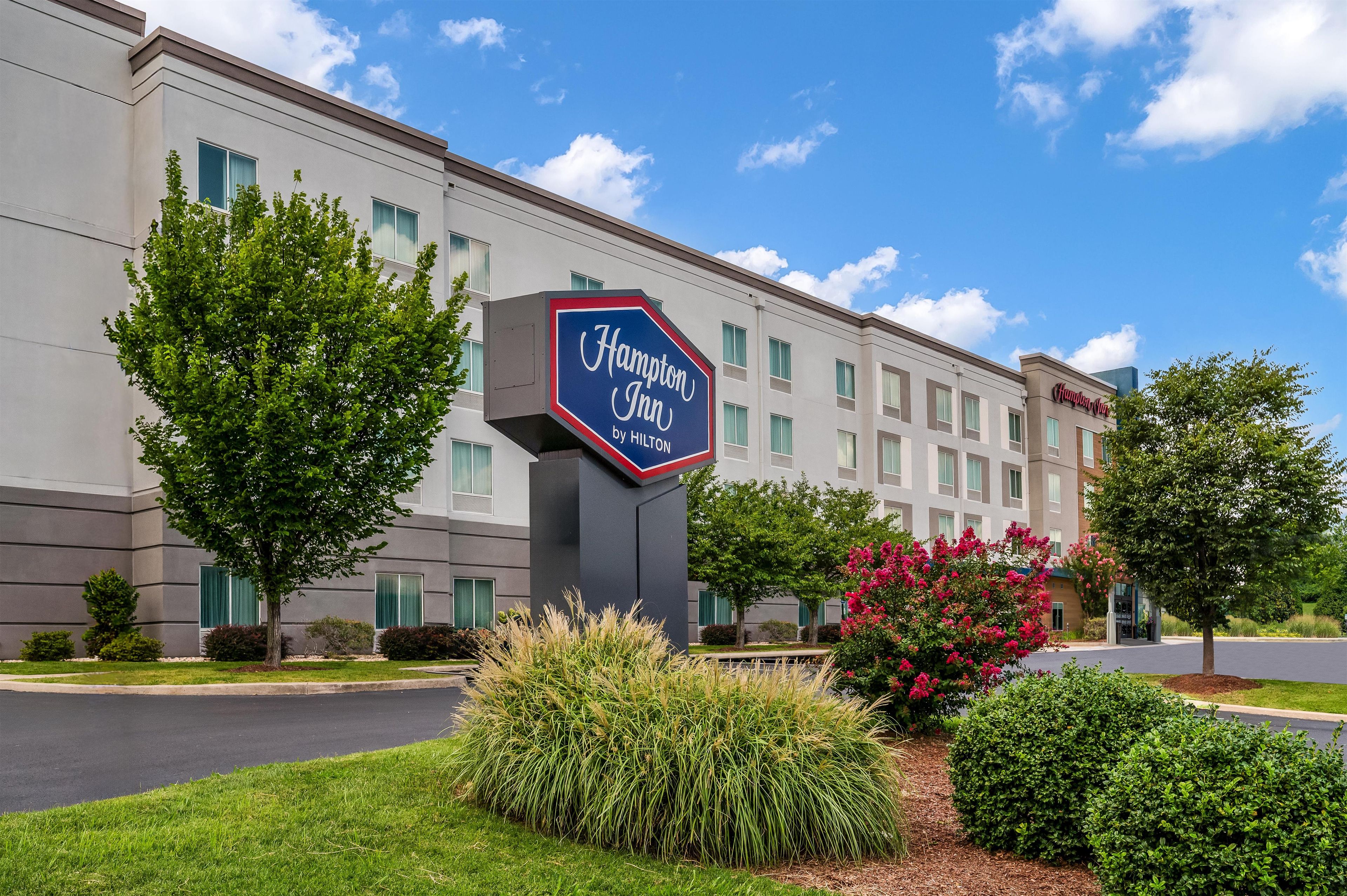 A Hampton Inn by Hilton hotel with a large sign, surrounded by well-maintained landscaping and trees.