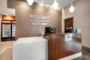 A reception desk with a 'WELCOME THE VOLUNTEER STATE' sign, a minibar, and a 'Hilton Honors' loyalty program sign. - Hampton Inn Nashville Airport Century Place, TN (Nashville)