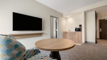 A hotel room with a flat-screen TV mounted on the wall, a round wooden table, a patterned armchair, and a kitchenette area with a microwave and coffee maker.