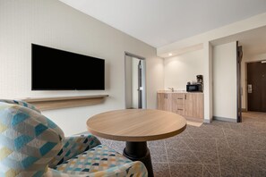 A hotel room with a flat-screen TV mounted on the wall, a round wooden table, a patterned armchair, and a kitchenette area with a microwave and coffee maker. - Hampton Inn Nashville Airport Century Place, TN (Nashville)