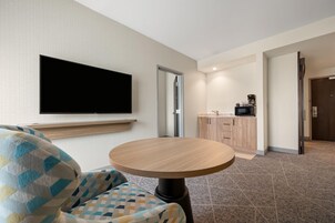 Habitación de hotel con una televisión de pantalla plana montada en la pared, una mesa redonda de madera, un sillón estampado y una zona de kitchenette con microondas y cafetera.