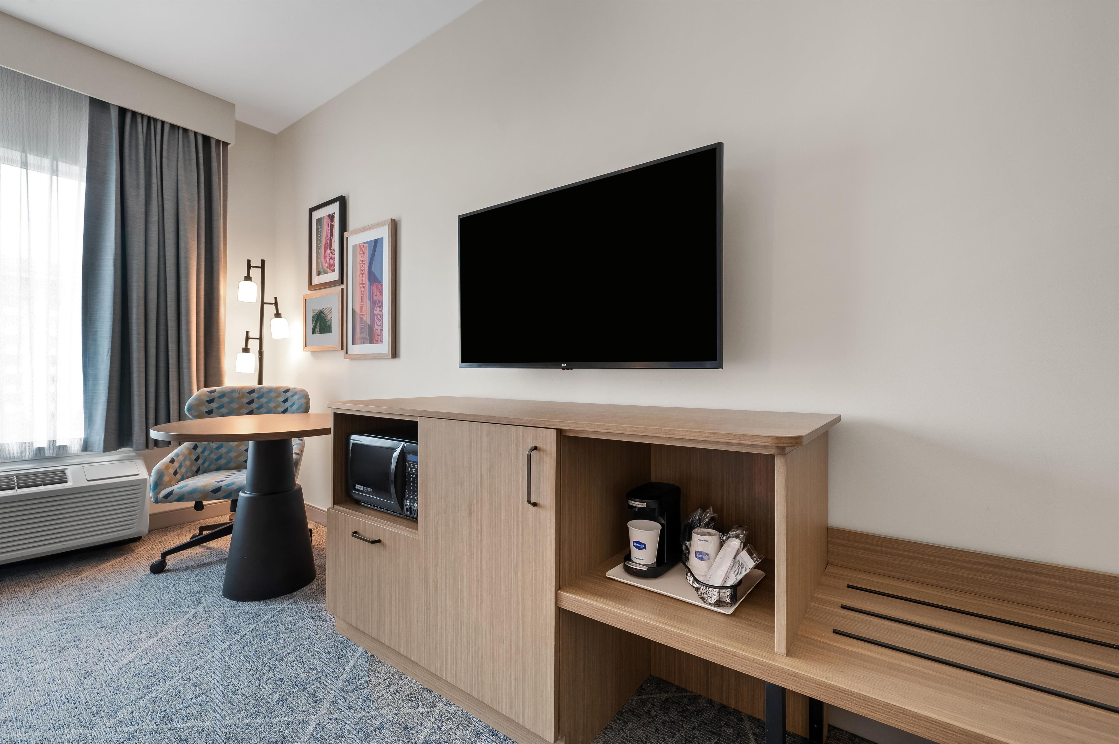 Habitación de hotel con televisor de pantalla plana en la pared, kitchenette con microondas, cafetera y una zona de estar con una silla y una mesita.