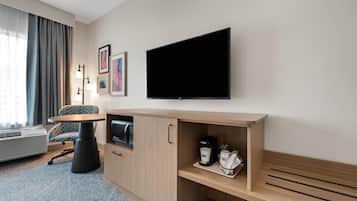 Habitación de hotel con televisor de pantalla plana en la pared, kitchenette con microondas, cafetera y una zona de estar con una silla y una mesita.