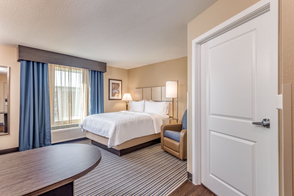 A hotel room with a bed, a desk, a chair, a lamp, and a door.