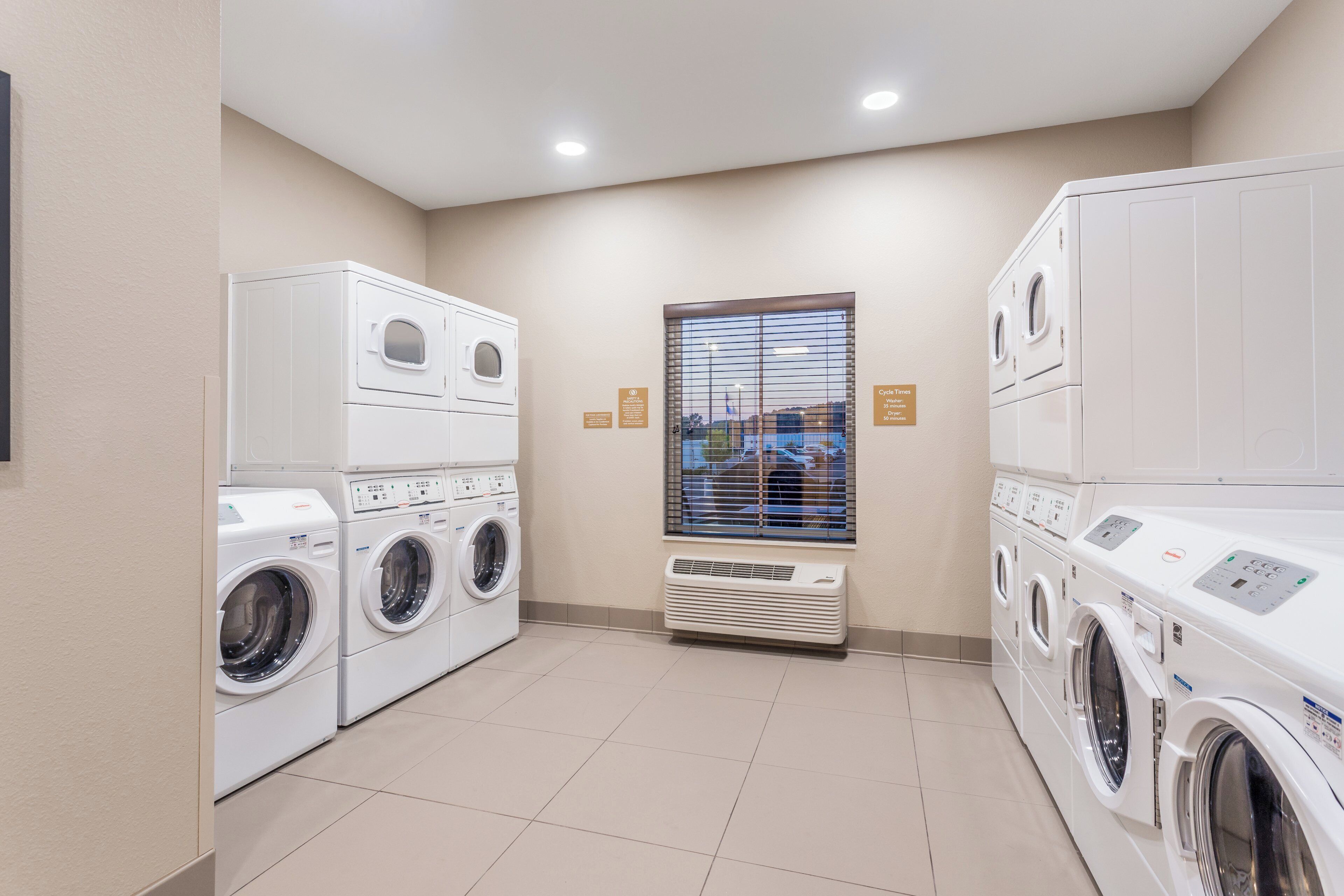 A laundry room with multiple washing machines and dryers, a window with blinds, and a ceiling with recessed lighting.