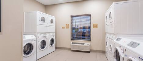 A laundry room with multiple washing machines and dryers, a window with blinds, and a ceiling with recessed lighting.