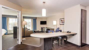 A hotel room with a kitchenette, a bed, a sitting area with a sofa and a lamp, a flat-screen TV, and a large window with curtains.
