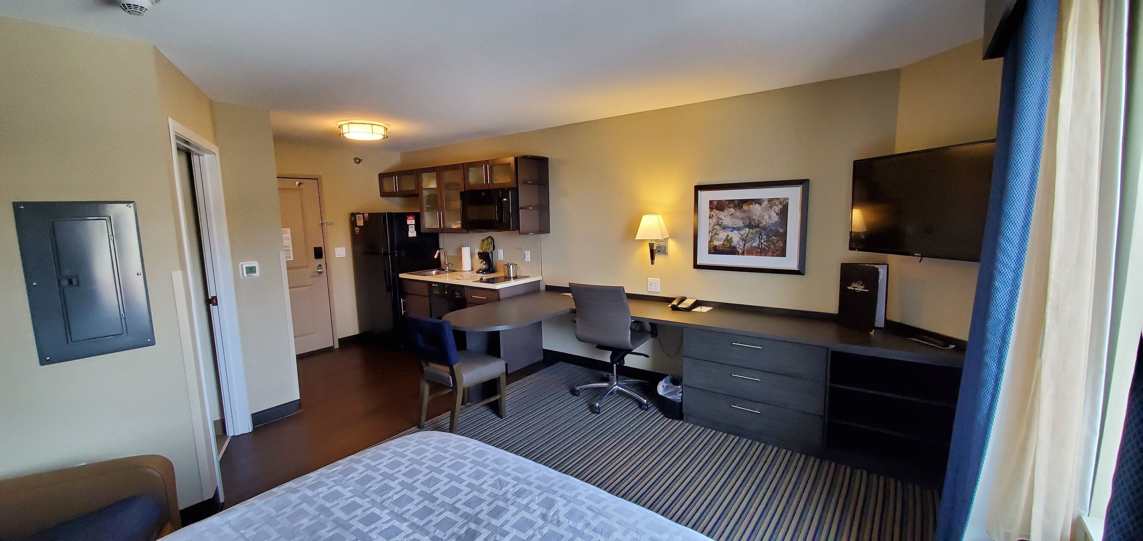 Une chambre d’hôtel comprenant un lit, un bureau, une chaise, une télévision, un coin cuisine et une fenêtre avec des rideaux.