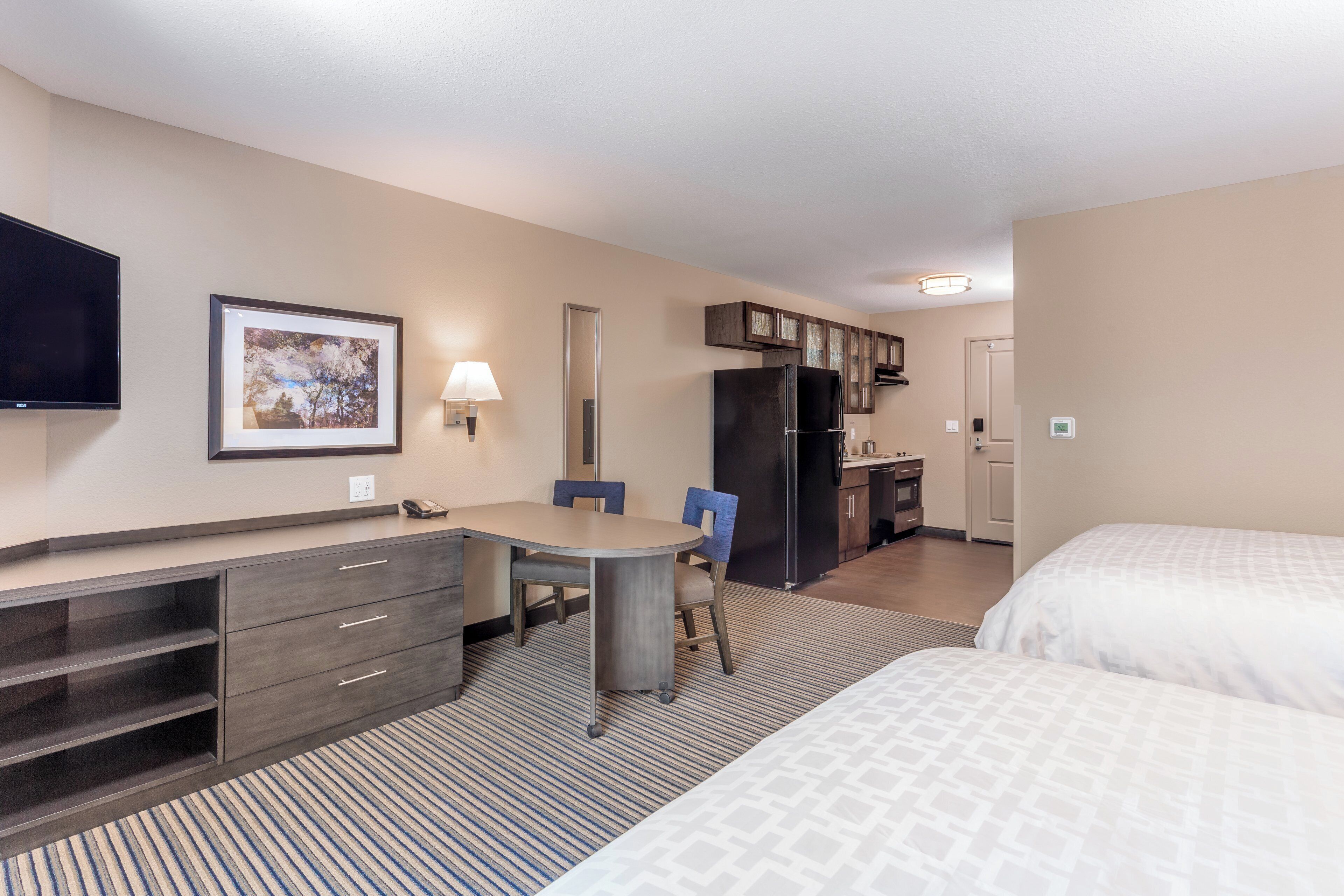 Una habitación de hotel con cama, escritorio con silla, kitchenette pequeño y televisor en la pared.