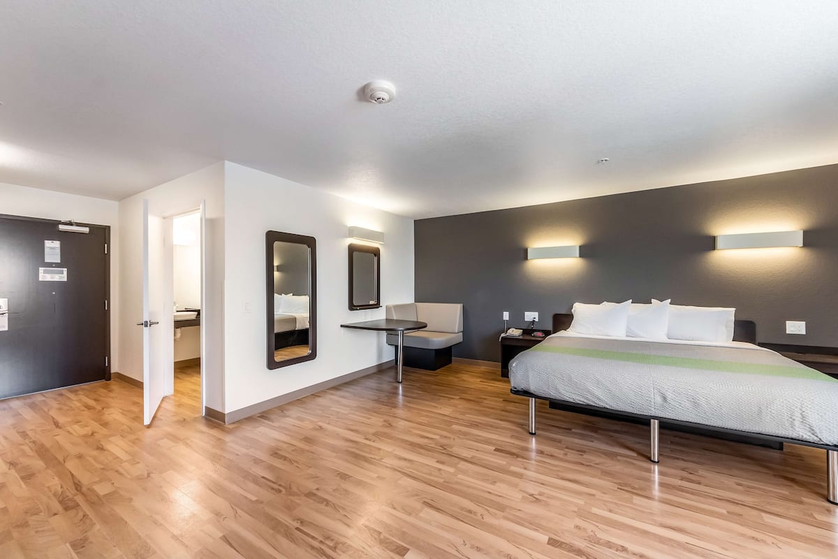 A modern hotel room with a wooden floor, a bed with white and green bedding, a small table, a chair, and two wall-mounted lights.