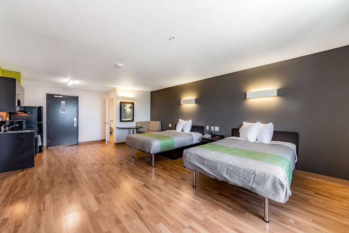 A modern hotel room with two beds, a kitchenette, and a bathroom.