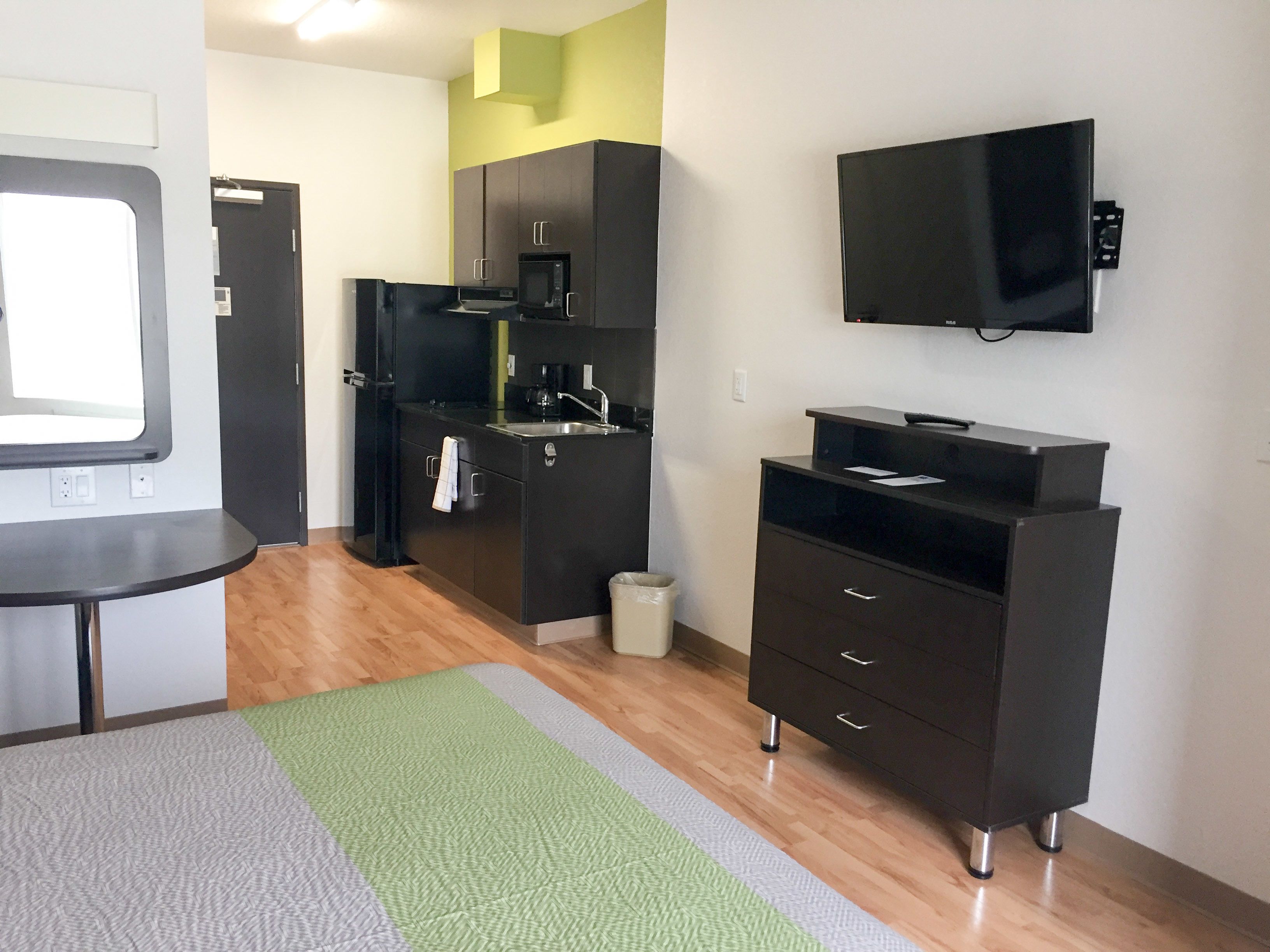 A compact kitchen with a microwave, sink, and dark cabinetry. There's a small trash bin and a flat-screen TV mounted on the wall.